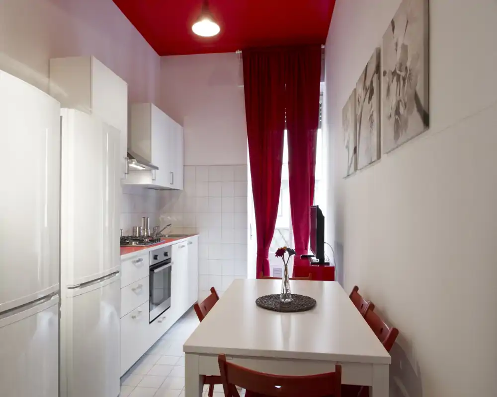 Bright, modern kitchenette with white cabinets, full-height fridge, built-in oven and a small dining table dressed with red accents and a flower vase.