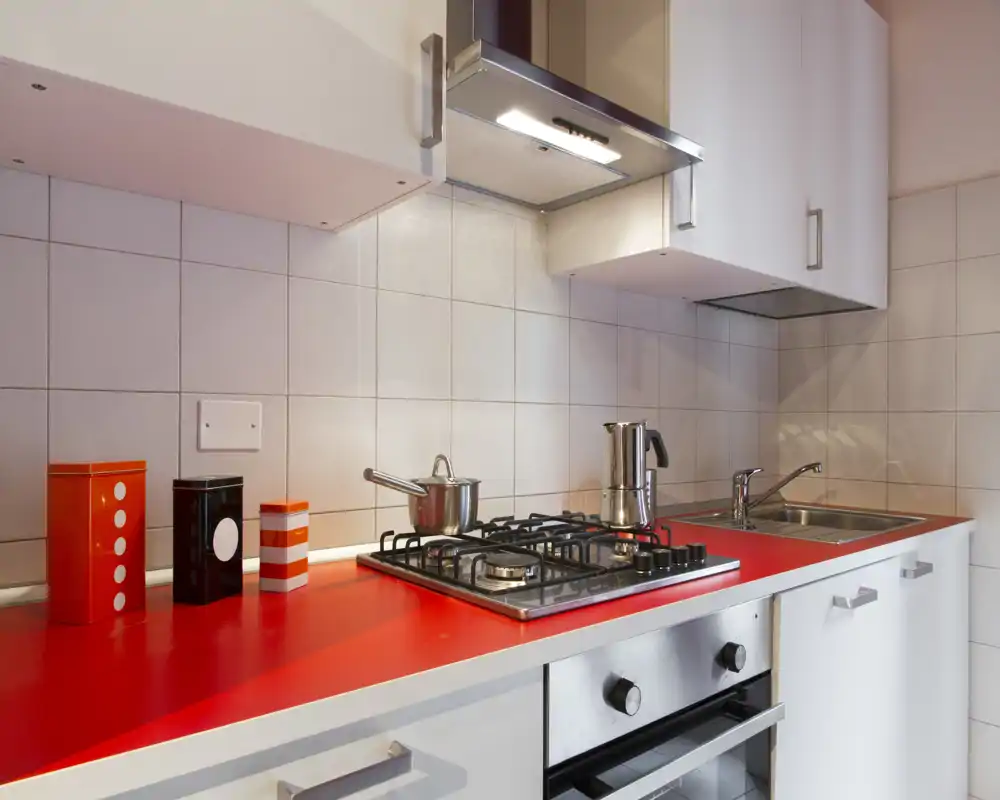 Modern compact kitchen with a red countertop, gas hob, stainless steel sink and overhead extractor — neat styling with colorful canisters adds personality.