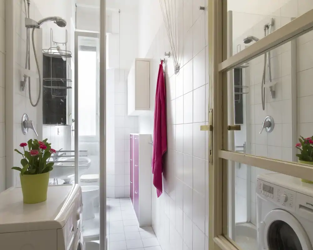 Bright, compact bathroom with a shower enclosure, washing machine and white tiled walls, accented by a pink towel and small potted plants.