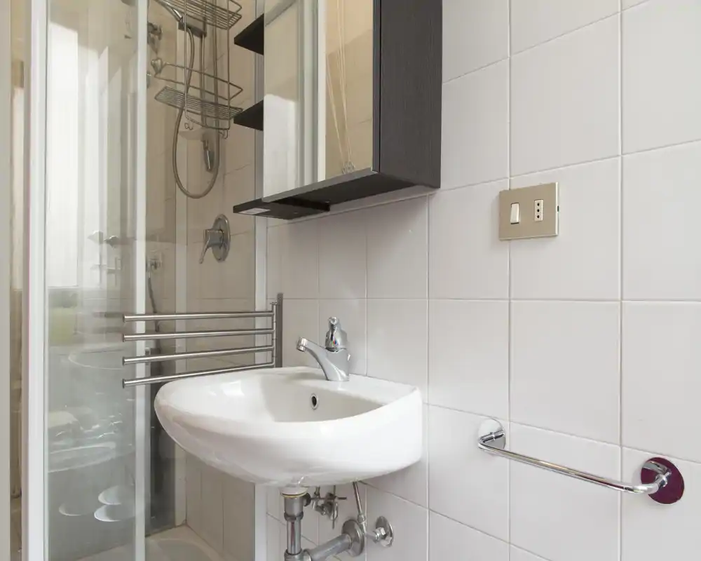 Clean, modern bathroom corner with a wall-mounted sink, chrome fixtures and a glass shower enclosure visible in the background.