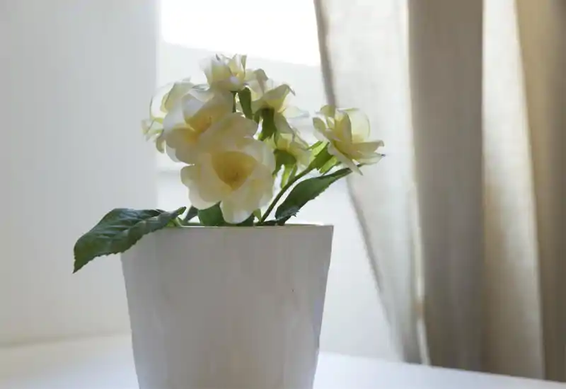 Close-up of a potted artificial-flower arrangement on a neutral table surface, lit by soft natural light near a window or curtain.
