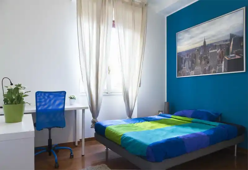 Dormitorio luminoso y ordenado con una cama doble colorida frente a una pared azul, ventana con cortinas translúcidas y un pequeño escritorio con silla azul — ideal para descansar y estudiar.