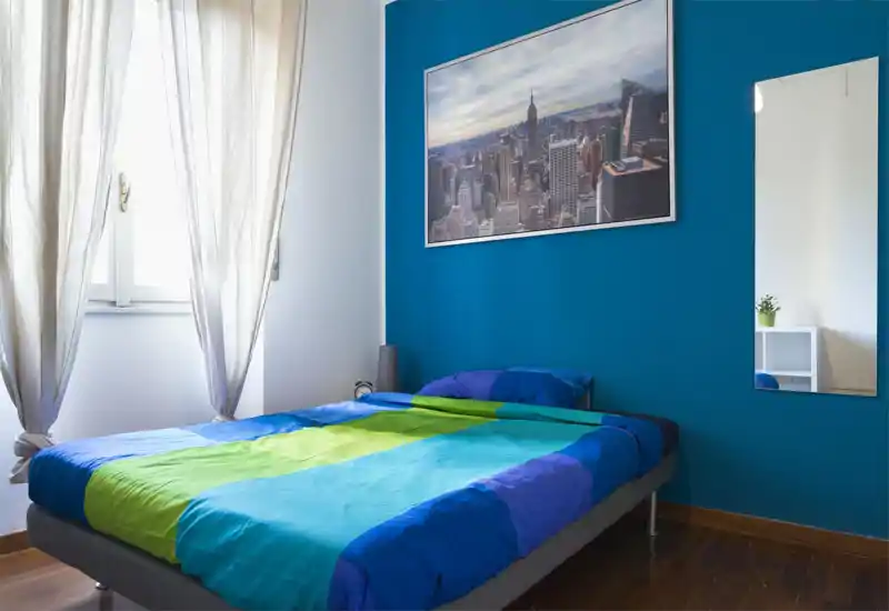 Dormitorio moderno y luminoso con cama doble de colores frente a una pared azul, gran ventana con cortinas claras y un espejo en la pared que aporta profundidad.