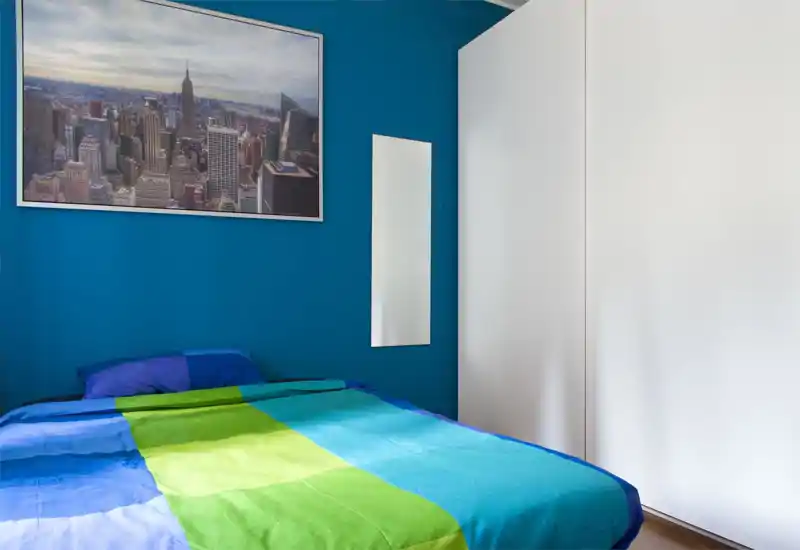 Dormitorio moderno y luminoso con cama doble de colores frente a una pared azul intensa y un gran armario blanco. Incluye una foto de la ciudad y un espejo estrecho; espacio ordenado y acogedor para estancias cortas o largas.