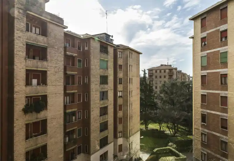 Bright city view from a window showing adjacent brick apartment buildings and a small green courtyard; good natural light and a pleasant urban outlook.