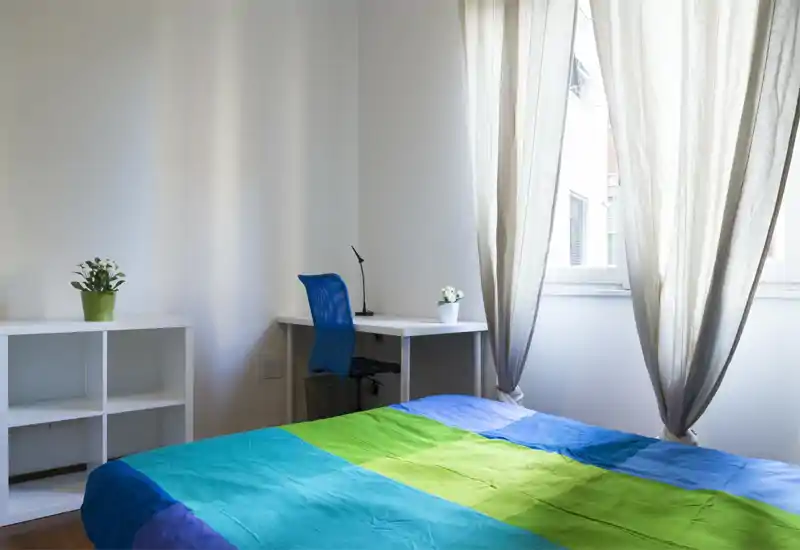 Dormitorio luminoso y minimalista con colcha colorida, escritorio y silla junto a la ventana, y cortinas que filtran la luz creando un ambiente tranquilo.