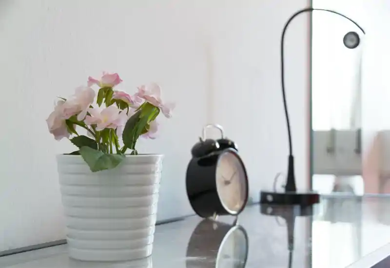 Una superficie de mesita luminosa con una maceta de flores rosas, un despertador negro clásico y una lámpara de lectura — un rincón de dormitorio tranquilo y minimalista.