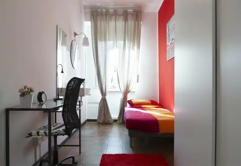 Dormitorio compacto y luminoso con cama individual de ropa de cama colorida junto a una ventana con cortinas ligeras; un pequeño escritorio y una silla ofrecen espacio de trabajo práctico.