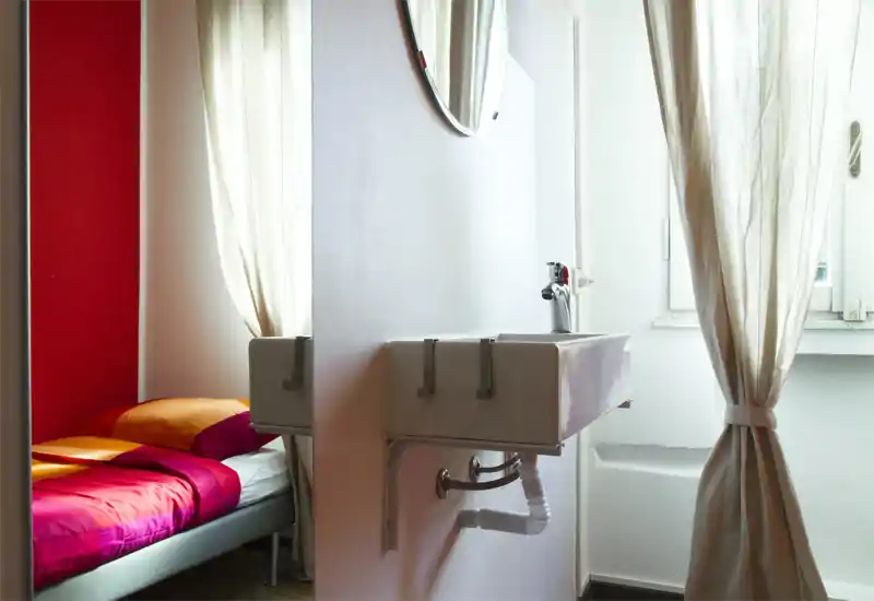 Dormitorio compacto con cama bien hecha en tonos naranja y magenta junto a un lavabo de pared; la luz natural que entra por las cortinas claras aporta una atmósfera acogedora.