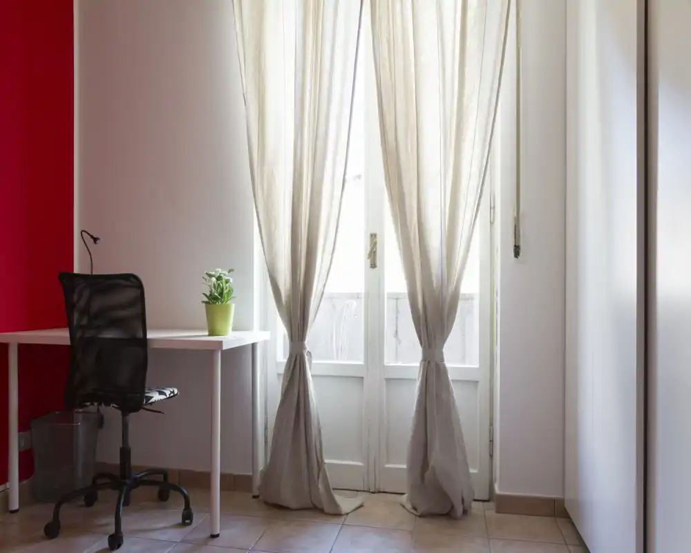 Espacio luminoso y minimalista con escritorio y silla de oficina junto a una puerta-finestra con cortinas de lino claro — ideal como pequeña oficina en casa o rincón de estudio.