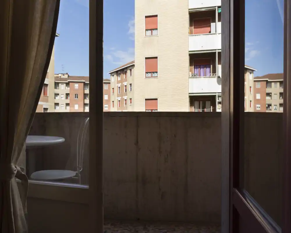 Pequeño balcón con barandilla de hormigón y una mesa y silla compactas, con vistas a bloques de apartamentos cercanos bajo un cielo despejado.