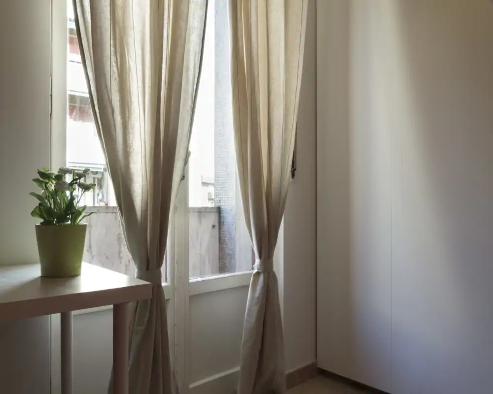 Una puerta con cortinas beige claro recogidas que dejan ver un pequeño balcón. Una planta en maceta sobre una mesa simple crea un rincón tranquilo y luminoso.