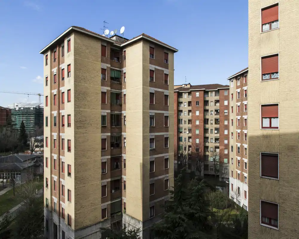 Vista exterior de bloques de apartamentos residenciales con fachadas de ladrillo y patios, fotografiada a plena luz del día. Ideal para mostrar el estado del edificio, las persianas y la vegetación exterior.