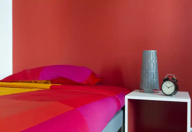 Dormitorio colorido con pared roja intensa y cama bien hecha en tonos magenta y rojo, además de una mesita con lámpara y despertador.