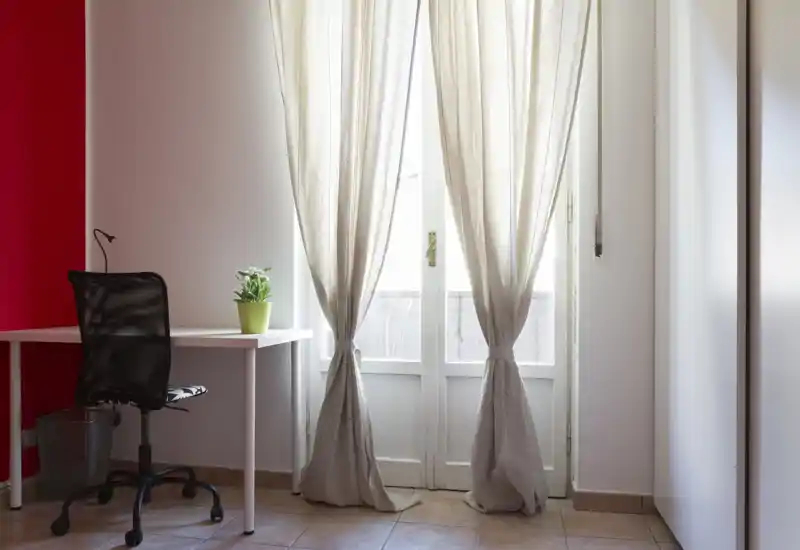 Espacio luminoso y minimalista con escritorio y silla de oficina junto a una puerta-finestra con cortinas de lino claro — ideal como pequeña oficina en casa o rincón de estudio.
