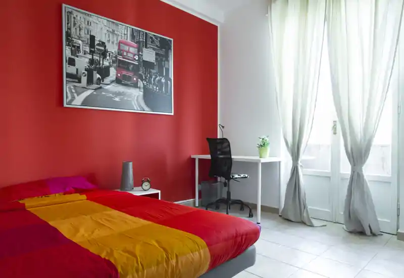 Dormitorio luminoso y colorido con una cama grande en primer plano, pared roja con cuadro, un escritorio y silla junto a la ventana soleada — ideal para anuncios que destaquen la comodidad y la luz natural.