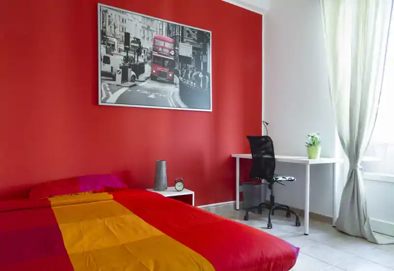 Dormitorio moderno y luminoso con una cama colorida frente a una pared roja, mesita con despertador y un escritorio sencillo junto a la ventana con luz natural.