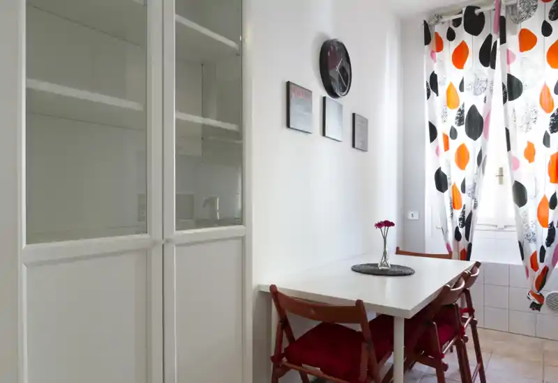 Compact dining nook with a white table, red folding chairs and a bright patterned curtain; natural light from the window creates a cheerful, well-styled corner ideal for small meals.