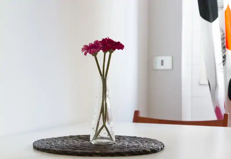 Minimal dining corner with a simple white table, a woven placemat and a clear glass vase holding pink flowers — bright, airy and tastefully minimalist.