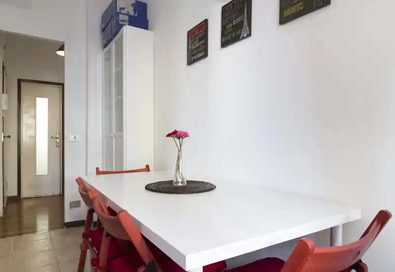 Bright, modern dining area with a white rectangular table, red chairs and a simple centerpiece vase with flowers — ideal for casual meals and compact spaces.
