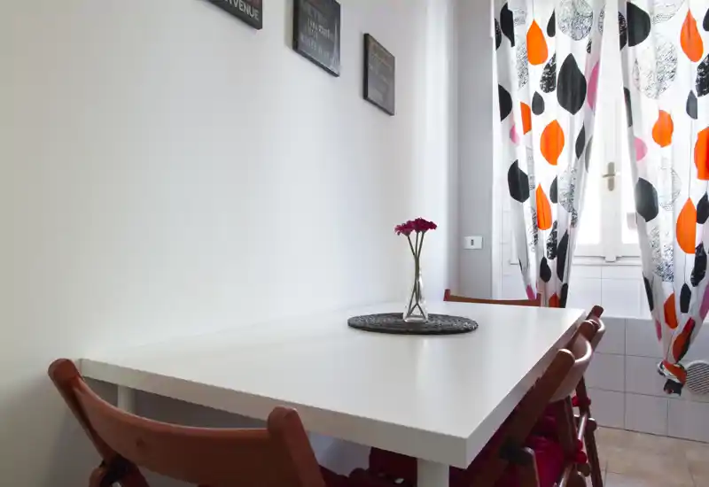 Bright dining nook with a simple white table, red chairs and patterned curtains; a small vase and placemat add a cozy touch.