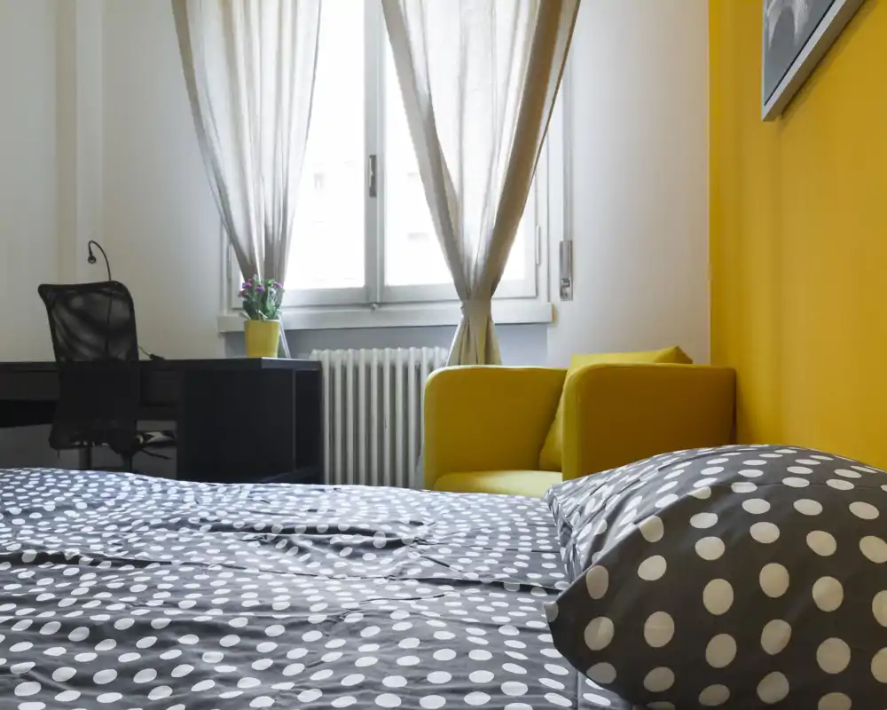 Bright, modern bedroom with polka-dot bedding, a yellow accent wall and a cozy armchair by a window letting in natural light.