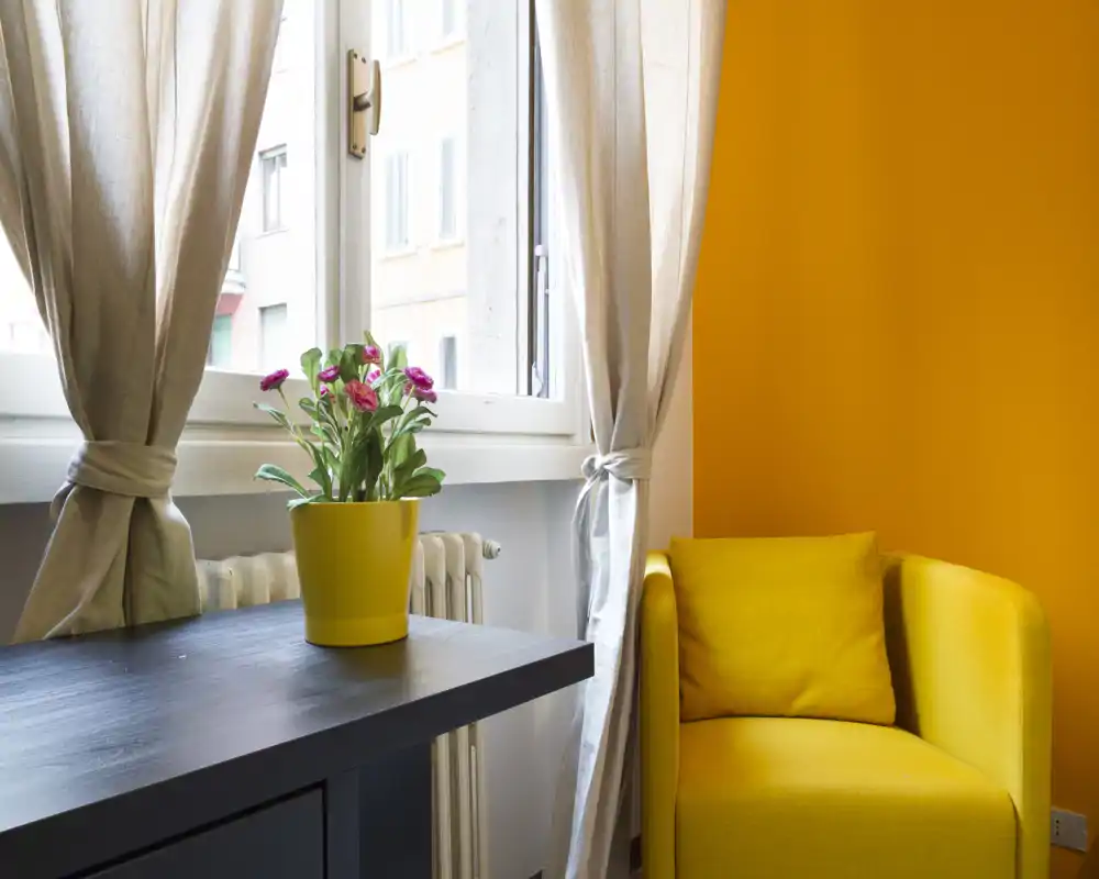 Bright, cozy corner of a living area with a yellow armchair, decorative cushion, a small table and a potted plant by the window, ideal for reading or relaxing.