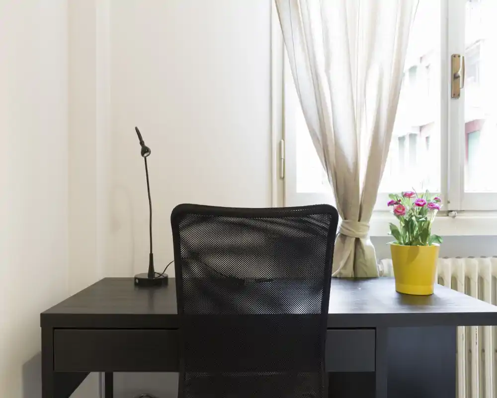Bright, minimalist workspace with a dark desk, mesh office chair, flexible desk lamp and a small potted plant by the window — ideal for a focused home office corner.