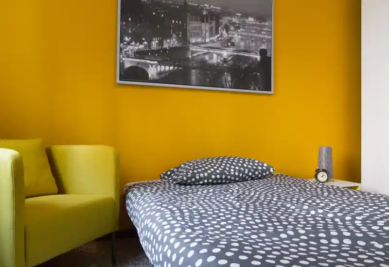 Camera da letto moderna e luminosa con copriletto a pois e una comoda poltrona davanti a una parete arancione, perfetta per un soggiorno accogliente.