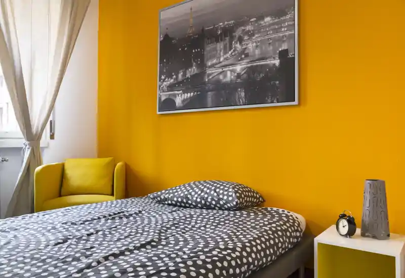 Camera da letto luminosa e accogliente con un letto matrimoniale a fantasia davanti a una parete arancione vivace, una comoda poltrona gialla vicino alla finestra e un comodino minimal con lampada e sveglia.