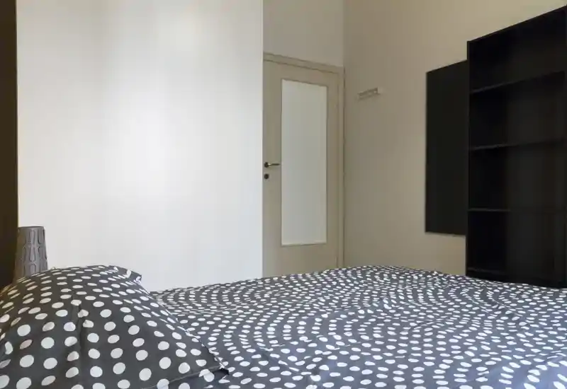 Camera da letto luminosa e minimal con copriletto a pois, lampada da comodino e mobile a scaffali — pareti neutre e linee pulite per un ambiente rilassante.