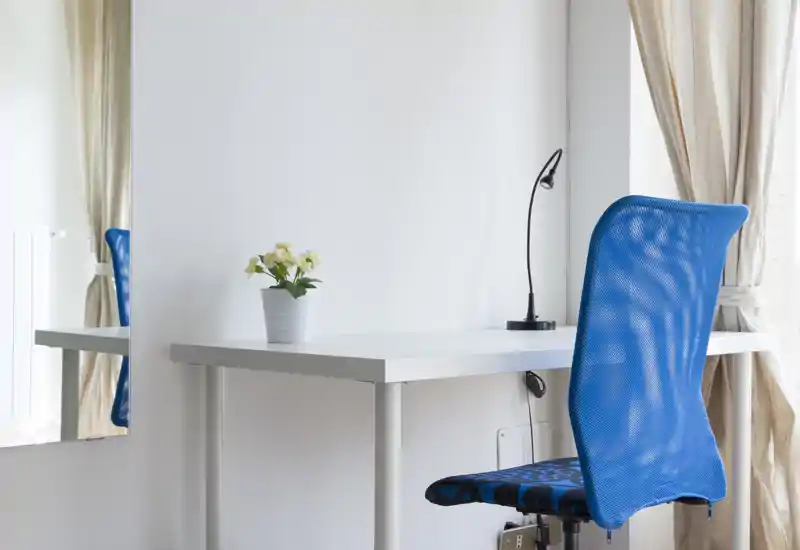 Angolo studio luminoso e minimale con scrivania bianca, lampada da tavolo flessibile, una piccola pianta in vaso e una sedia ergonomica blu accanto alla finestra con tende.