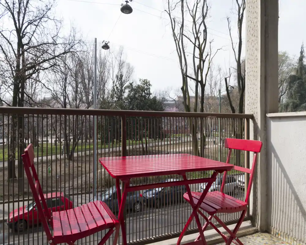 Balcone soleggiato con un piccolo tavolo bistro rosso e due sedie abbinate che si affacciano su una strada alberata — ideale per un caffè mattutino o una pausa all'aperto.