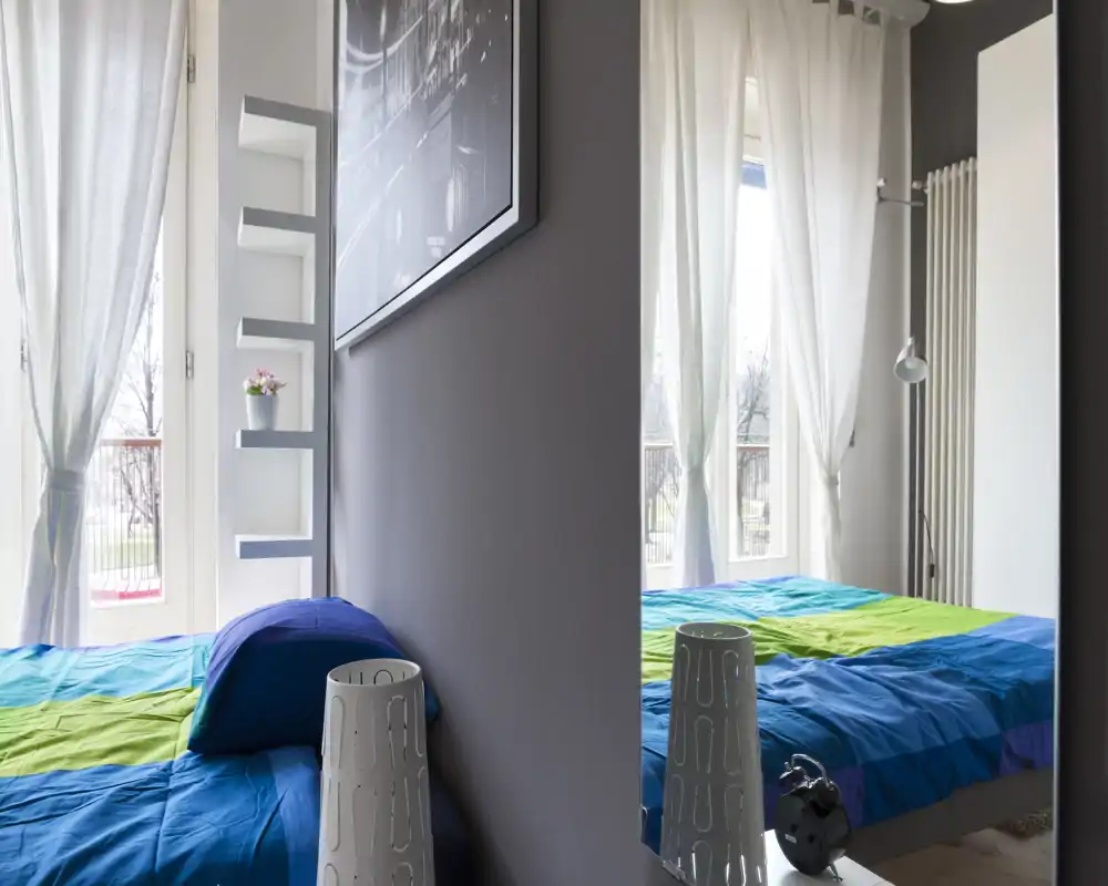 Camera da letto moderna e luminosa con una trapunta blu e verde, grandi finestre con tende leggere e dettagli decorativi che la rendono accogliente.