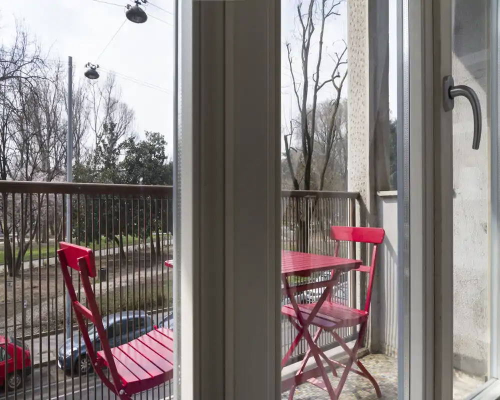 Balcone soleggiato con piccolo tavolino bistro rosso e due sedie abbinate, ringhiera in metallo e vista sulla strada alberata — ideale per il caffè mattutino.