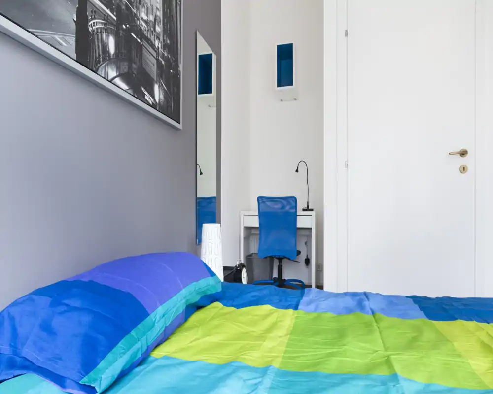 Camera da letto moderna e luminosa con biancheria colorata, piccola scrivania e sedia — ideale per una persona o uno studente.