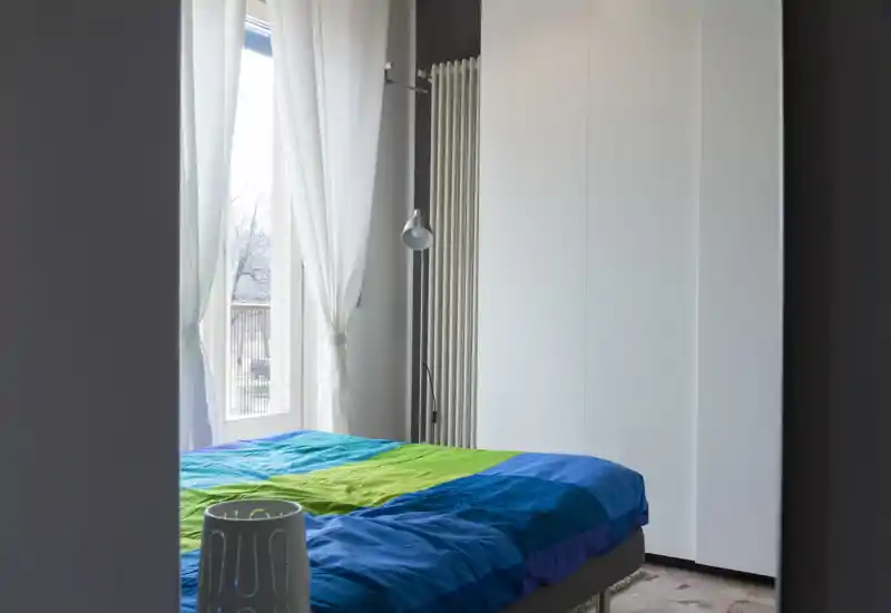 Camera da letto luminosa e ordinata con copriletto colorato, ampia armadiatura e tende leggere che filtrano la luce naturale — ideale per annunci che vogliono sottolineare comfort e spazio di stoccaggio.