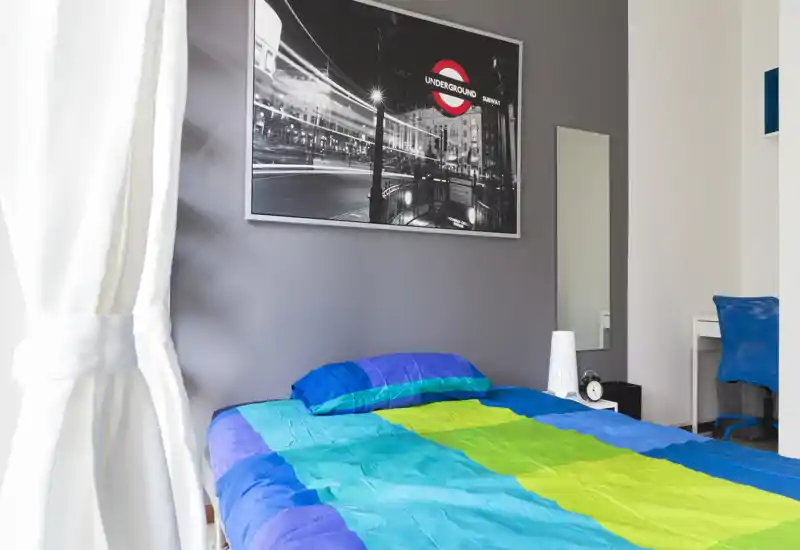 Camera da letto luminosa e moderna con un letto colorato, un piccolo comodino e una grande stampa appesa sopra la testata. La luce naturale dalla tenda a sinistra rende l'ambiente fresco e accogliente, perfetto per annunci di affitto.