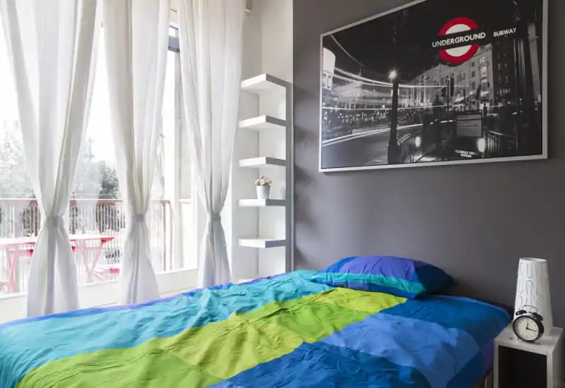 Camera da letto luminosa e moderna con un letto matrimoniale colorato accanto a una grande finestra e balcone; mensole minimal e quadro su parete donano carattere.