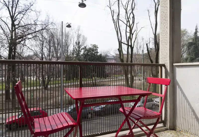 Balcone soleggiato con un piccolo tavolo bistro rosso e due sedie abbinate che si affacciano su una strada alberata — ideale per un caffè mattutino o una pausa all'aperto.