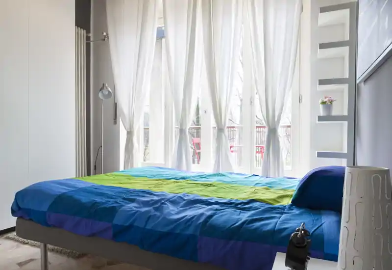 Camera da letto moderna e luminosa con un grande letto con copriletto colorato, abbondante luce naturale da finestre a tutta altezza e una piccola mensola decorativa.