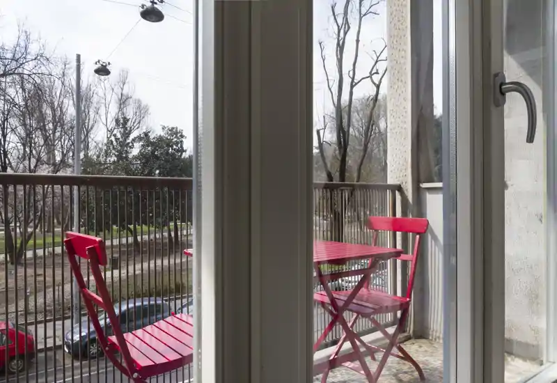 Balcone soleggiato con piccolo tavolino bistro rosso e due sedie abbinate, ringhiera in metallo e vista sulla strada alberata — ideale per il caffè mattutino.