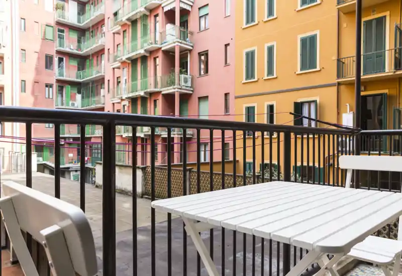 Balcone privato soleggiato con tavolo e sedie bianchi compatti che si affacciano su colorate facciate residenziali — ideale per un caffè mattutino o una pausa all'aperto.