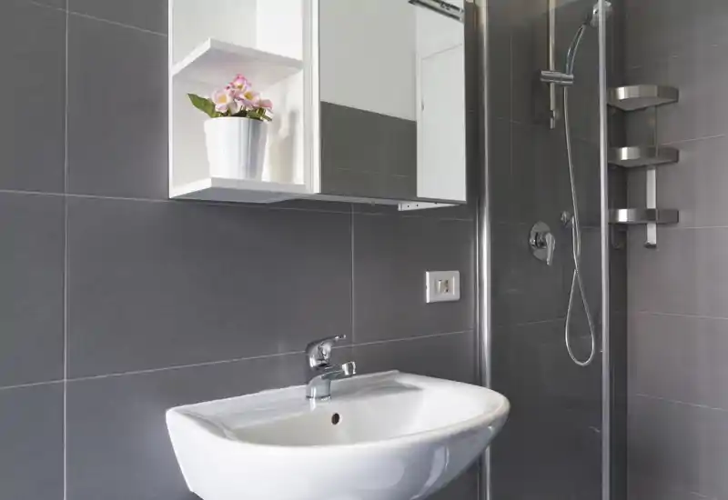 Bagno moderno piastrellato con lavabo a muro, specchiera contenitore e box doccia in vetro; ambiente pulito e ben illuminato con rubinetteria contemporanea.
