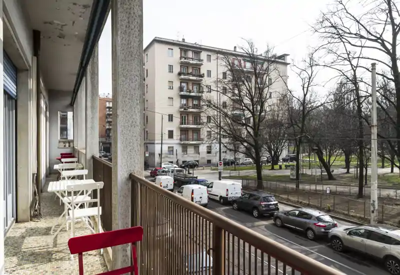 Balcone cittadino con tavolini e sedie pieghevoli che si affacciano su una strada alberata e palazzi residenziali — perfetto per il caffè mattutino.