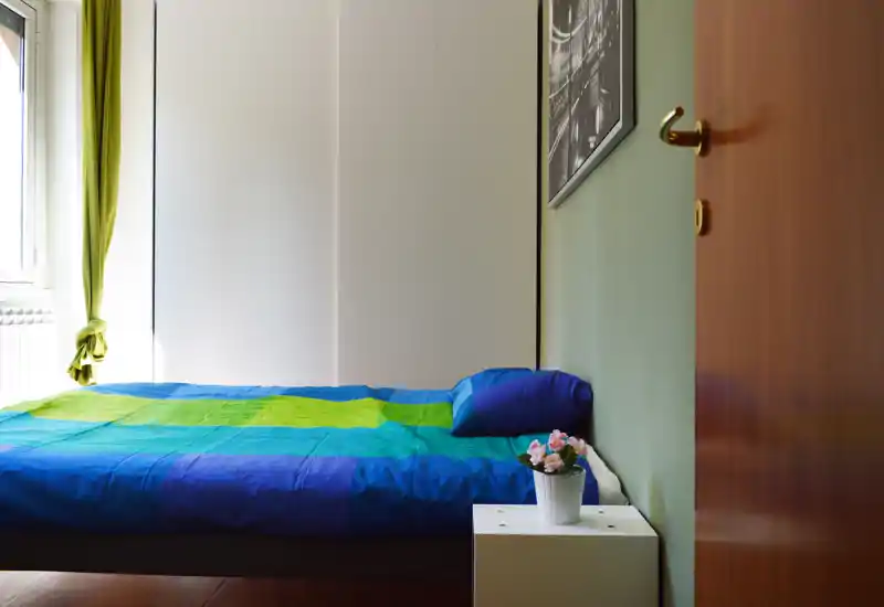 Dormitorio luminoso y ordenado con una cama doble con ropa de cama azul-verde, una almohada, una pequeña mesita blanca con una maceta con flores y luz natural desde la ventana.
