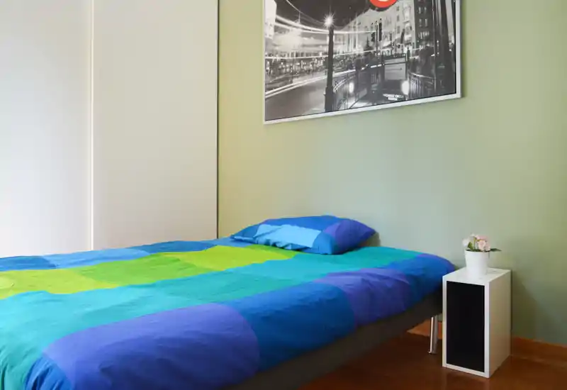 Dormitorio individual moderno y luminoso con ropa de cama colorida, una pequeña mesita y un cuadro en la pared: limpio, bien iluminado y listo para huéspedes.