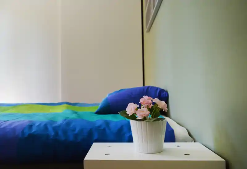 Rincón de dormitorio luminoso y ordenado con una cama azul y verde, una almohada junto a la pared y una maceta con flores sobre una mesita blanca.