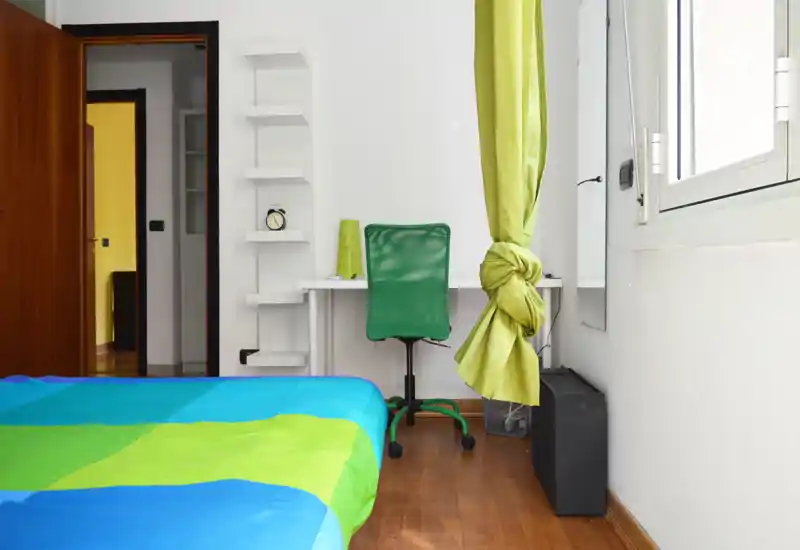 Dormitorio moderno y luminoso con colcha colorida, pequeño espacio de trabajo con silla verde y una gran ventana que aporta luz natural.