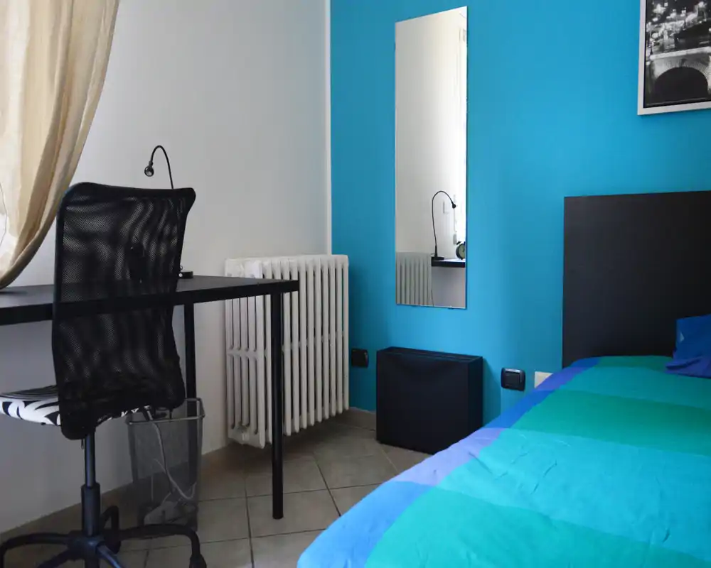 Pequeña habitación con cama individual con ropa azul-verde, cabecero negro y un escritorio compacto con silla de malla junto al radiador; pared turquesa y espejo de cuerpo entero que aportan color y profundidad.